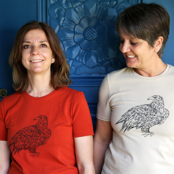 Zwei Frauen vor geschnitzter blauer Türe, sie tragen T-Shirtsin rot und beige mit Siebdruckmotiv Adler
