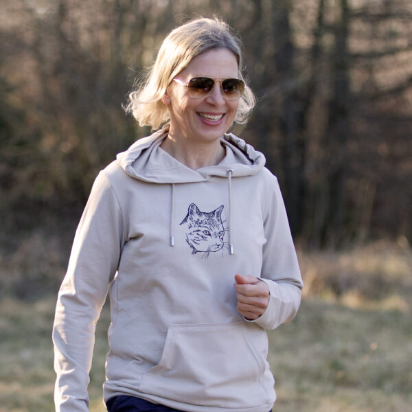 Lächelnde Frau bewegt sich in der Natur, sie trägt einen hellbraunen Hoodie mit Siebdruckmotiv Katzenkopf