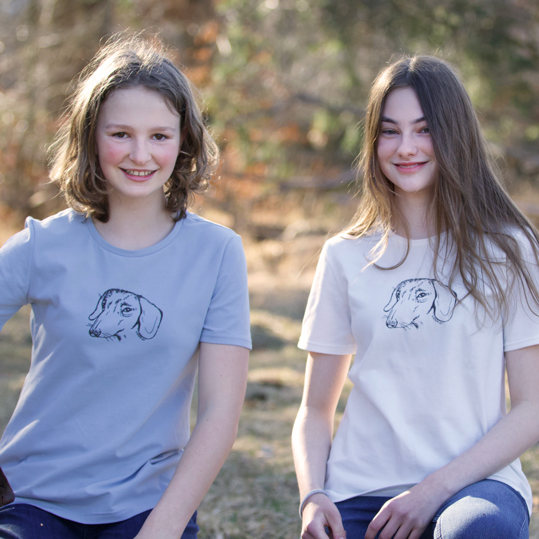Zwei junge Mädchen knien in der Natur, sie tragen Shirts, hellblau und weiß, jeweils mit Siebdruckmotiv Dackelkopf