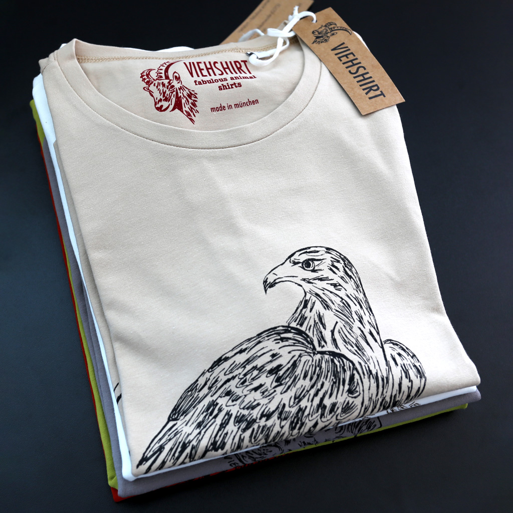 Stapel mit fünf zusammengelegten T-Shirts. Oben liegt ein hellbraunes T-Shirt mit Siebdruckmotiv Adler
