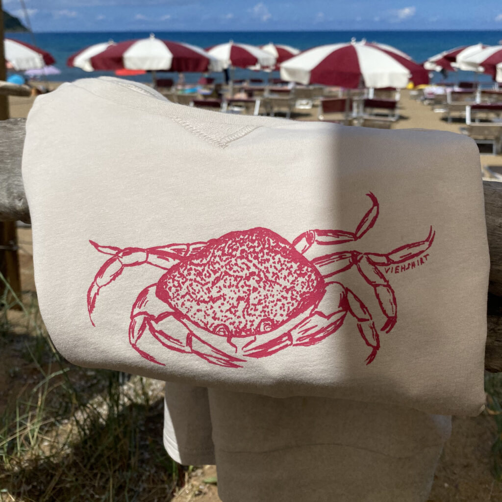 Sandfarbener Sweater vor Strandpromenade mit rotem Siebdruckmotiv Krabbe