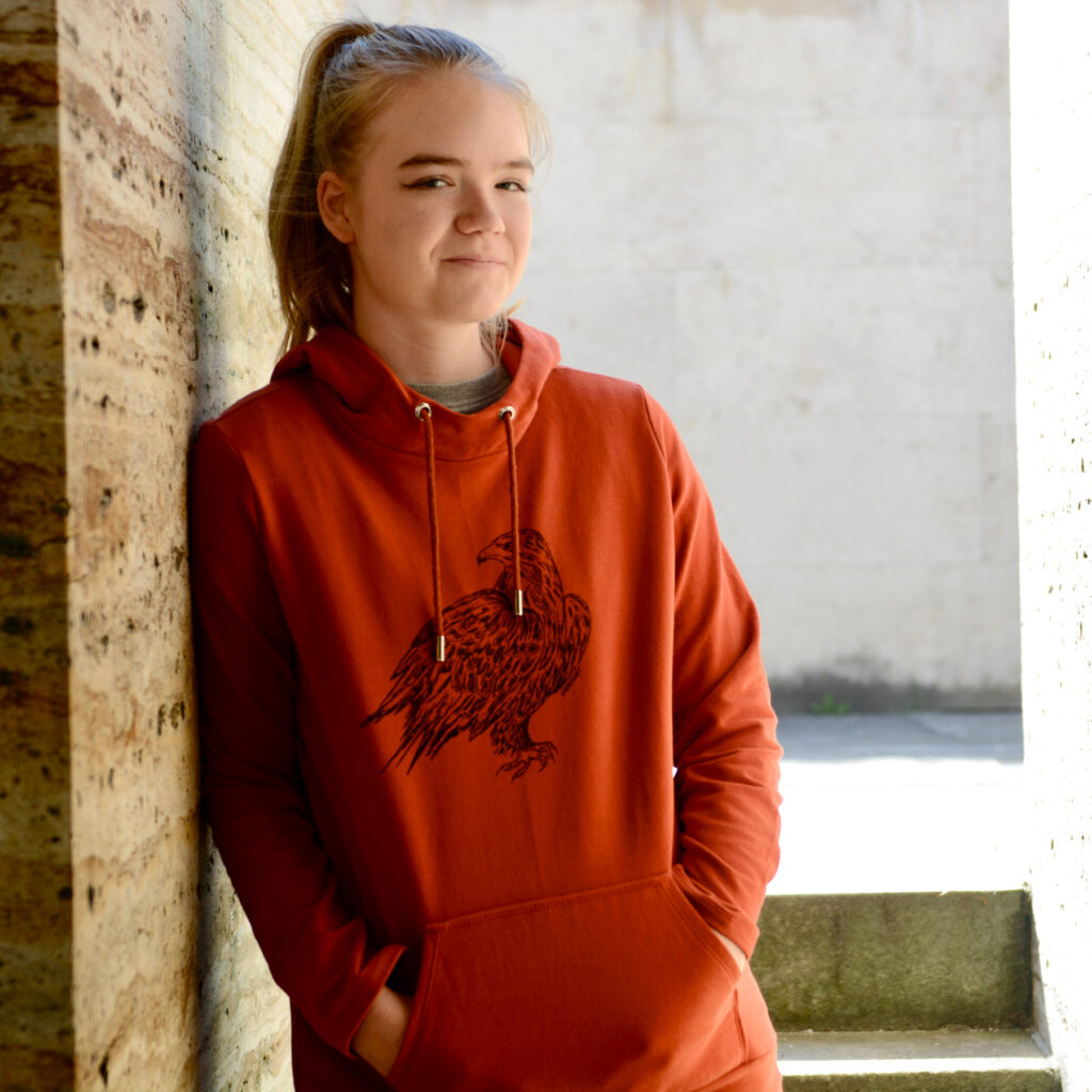 Junge Frau an Steinwand gelehnt trägt roten Hoodie mit Siebdruckmotiv Adler