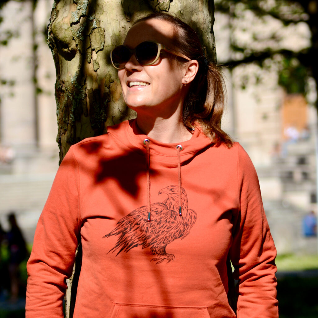 Frau mit Sonnenbrille an Baumstamm gelehnt, Lichterspiel, sie trägt roten Hoodie mit Siebdruckmotiv Adler