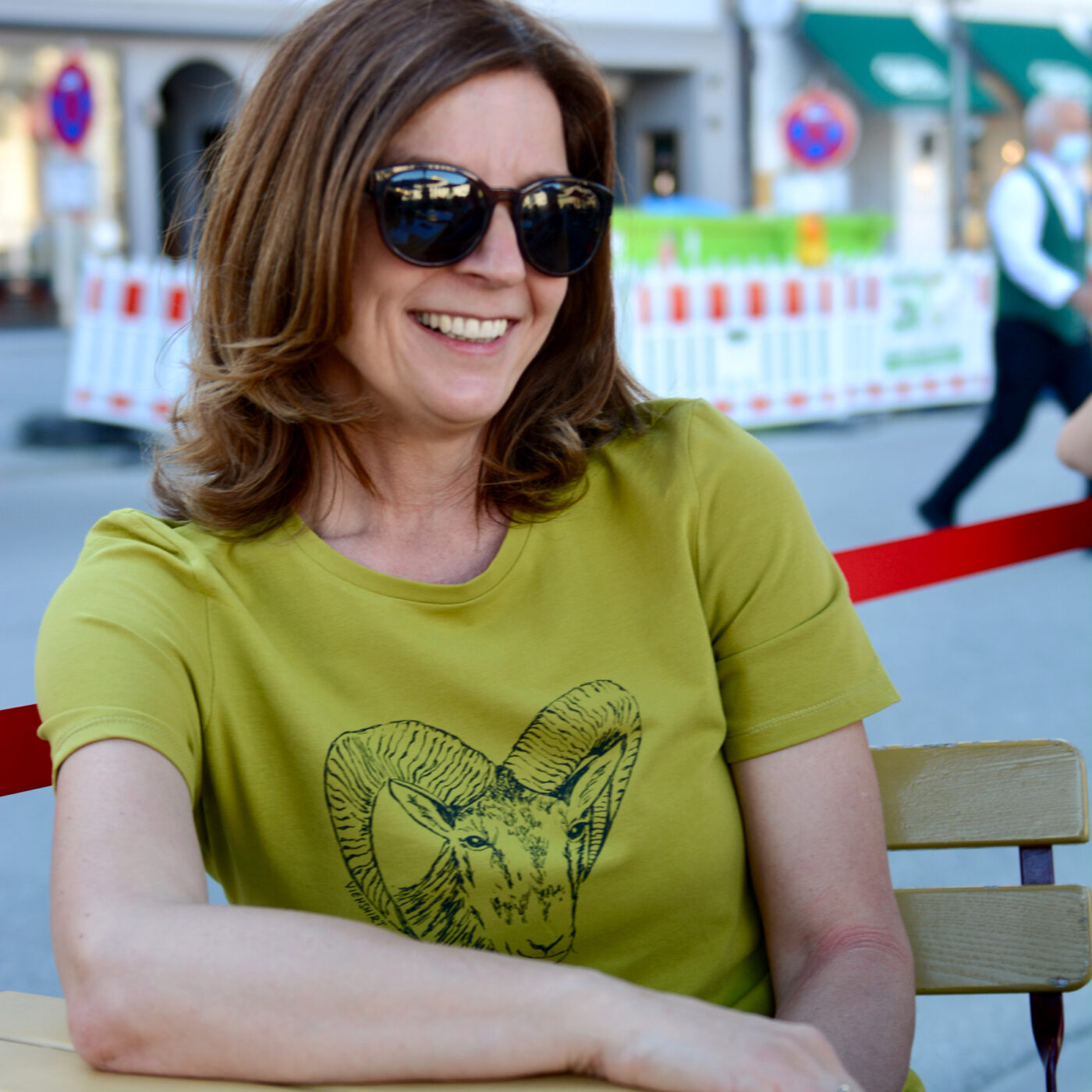 Frau vor städtischem Hintergrund, sie trägt ein grünes T-Shirt mit Siebdruckmotiv Mufflonkopf.