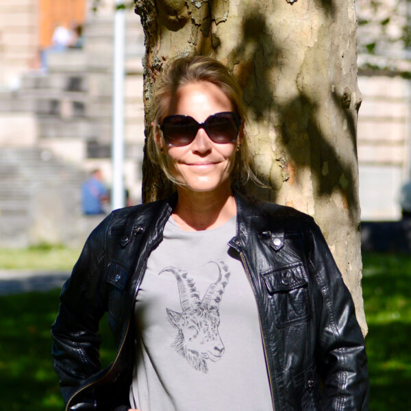 Frau mit Sonnenbrille an Baumstamm gelehnt, Lichterspiel, sie trägt Lederjacke und darunter ein graues T-Shirt mit Siebdruckmotiv Steinbock