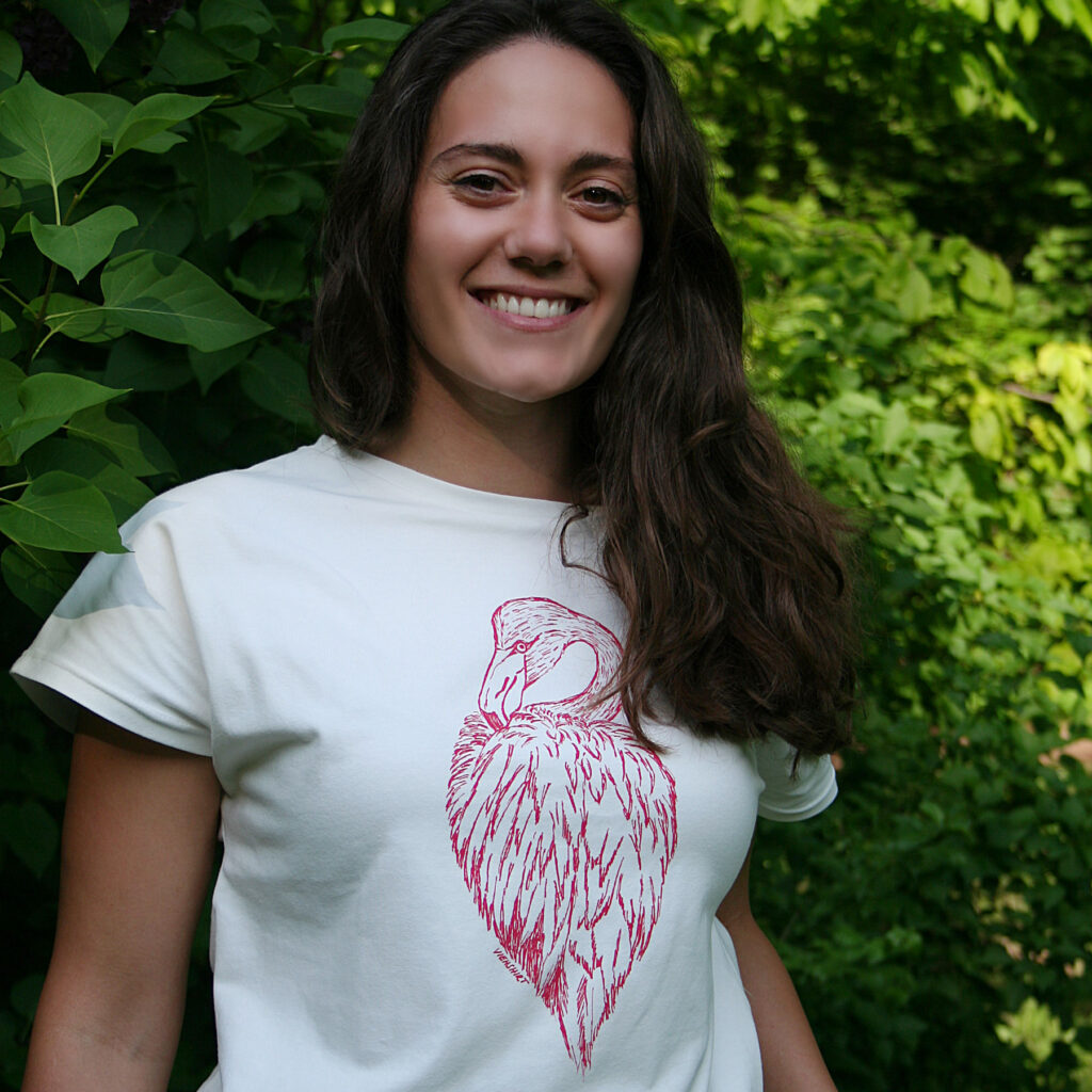 Frau mit langen braunen Haaren vor grünen Pflanzen trägt weißes Shirt mit Siebdruckmotiv Flamingo