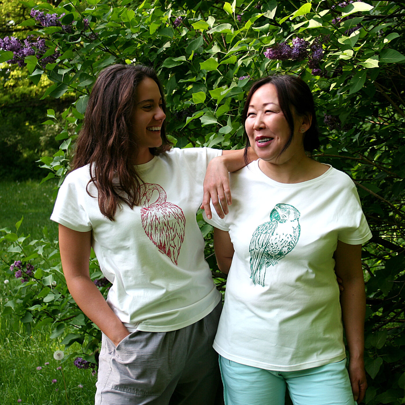 Zwei Frauen stehen vor grünen Pflanzen, sie tragen weißes Shirt mit Siebdruckmotiven Flamingo und Papagei
