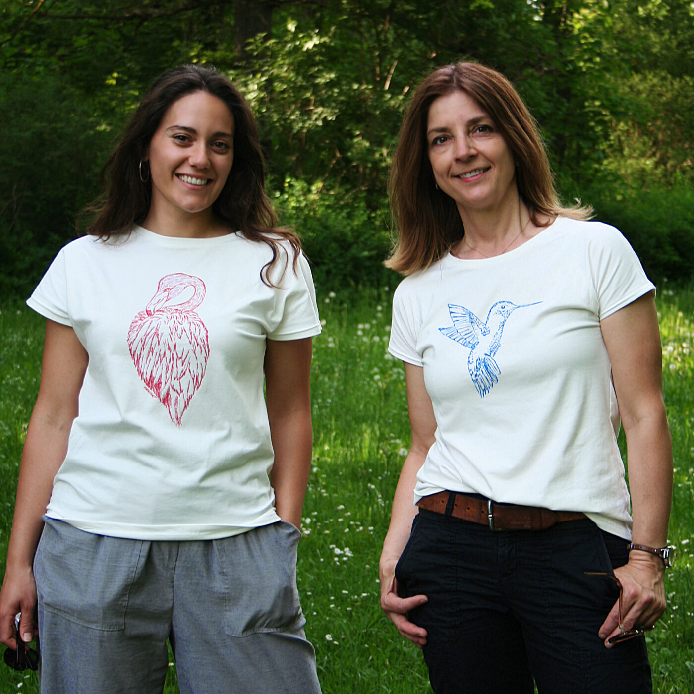 Zwei Frauen stehen vor grünen Pflanzen, sie tragen weißes Shirt mit Siebdruckmotiven Flamingo und Kolibri